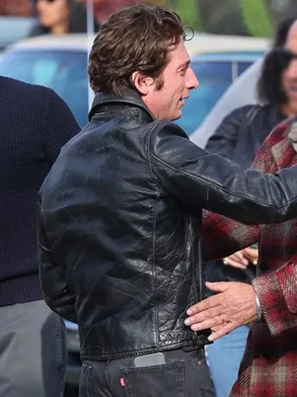 Man wearing a black leather jacket in an outdoor setting with blurred people and cars in the background