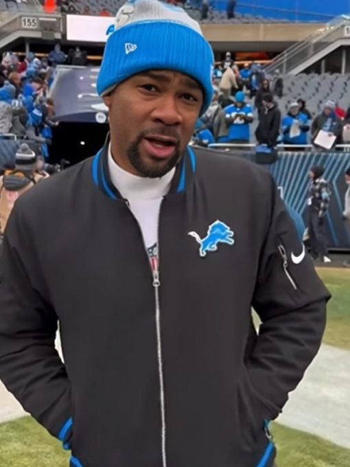 Person wearing a black jacket with a logo, blue beanie, and white shirt at a sports event.