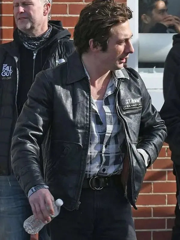 Man wearing a black leather jacket with a plaid shirt, standing against a brick wall.
