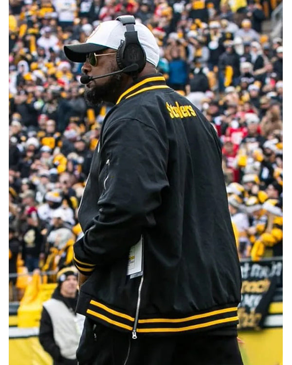 Person wearing a black jacket with yellow accents and a headset, standing in front of a crowd at a sports event.