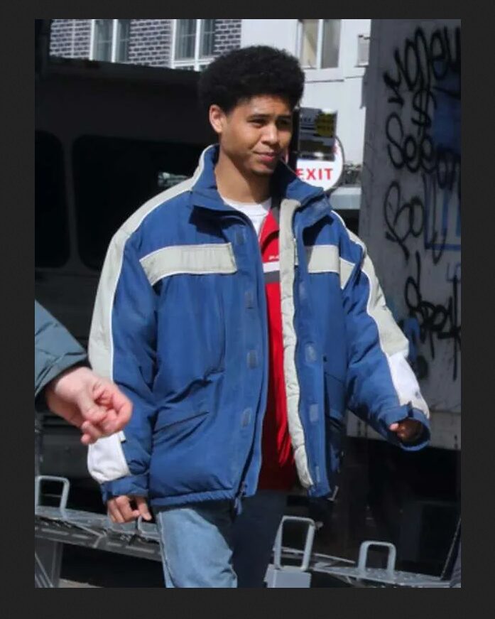 A person wearing a blue and white jacket with a stand-up collar, featuring a zipper and velcro closure, standing near a graffiti wall.